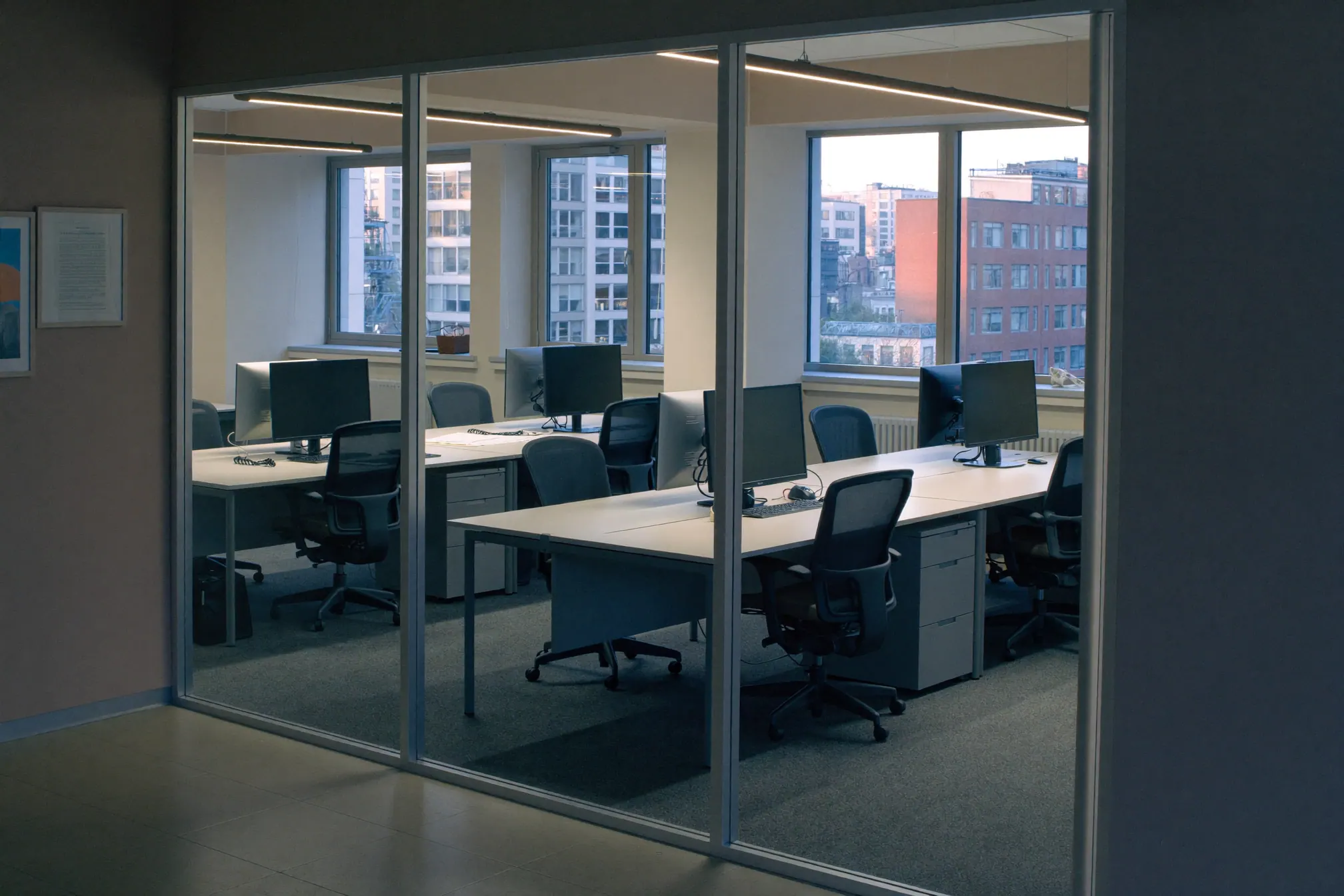 A small open-plan office seen through a glass partition at dawn, empty desks and monitors switched off