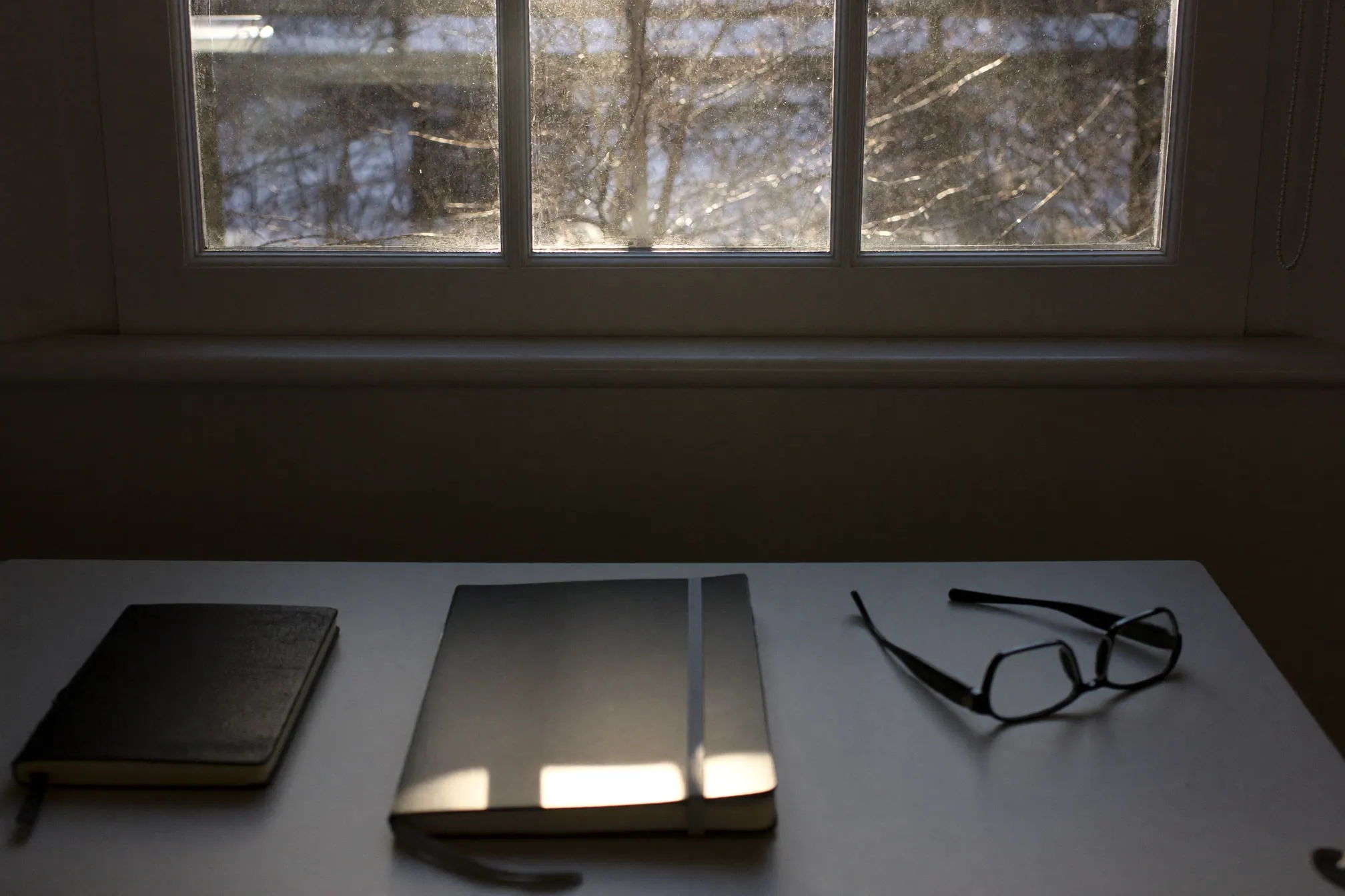 A desk by a window in late December with a closed notebook and a pair of glasses, low winter sun coming in
