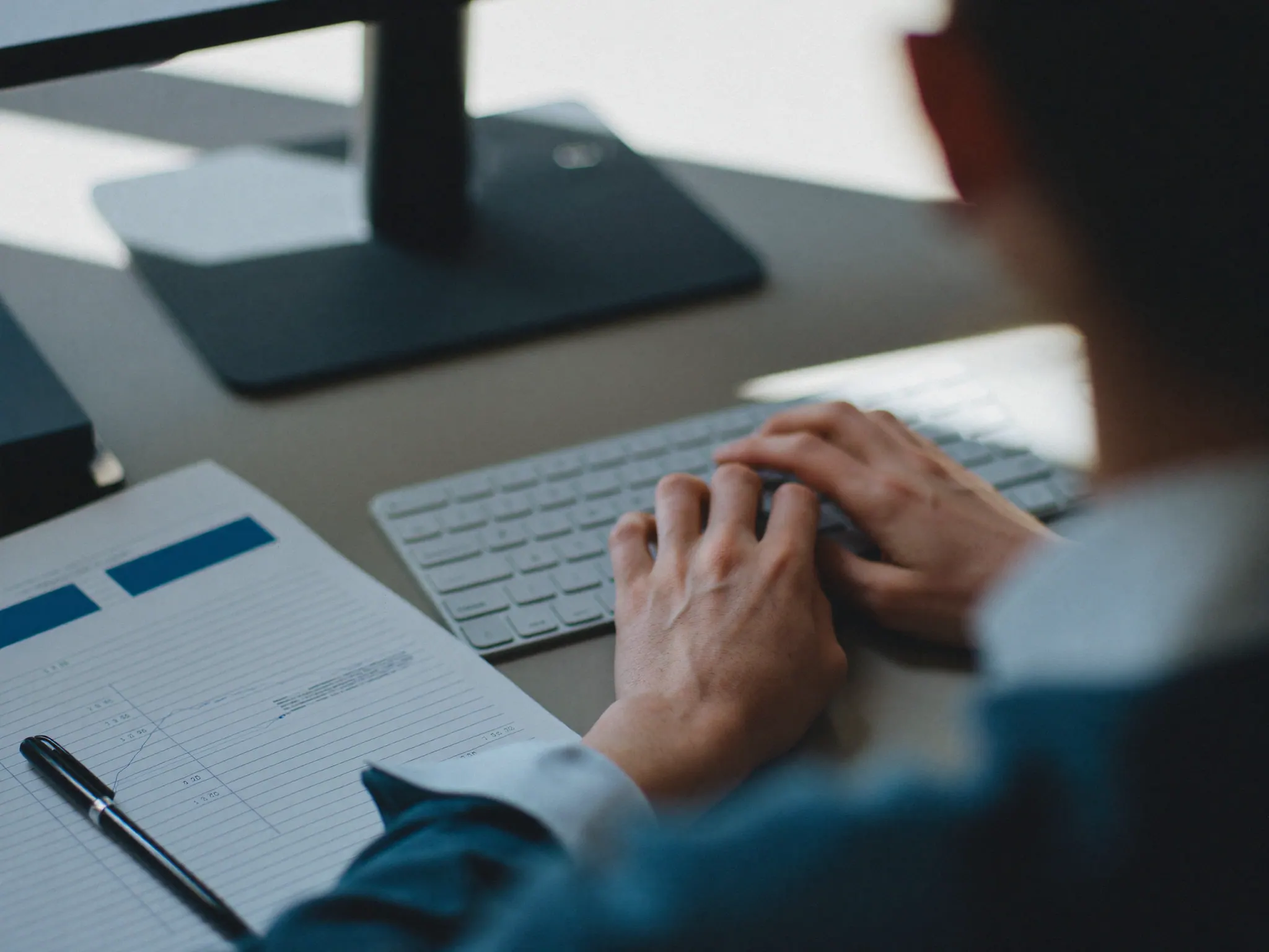 Over the shoulder of a person at a clean desk, hands on a keyboard beside a printed spreadsheet
