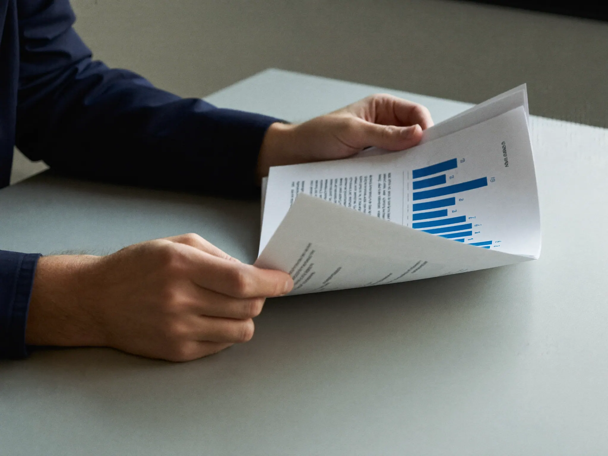 A person's hands turning the page of a printed report on a pale grey table in cool daylight