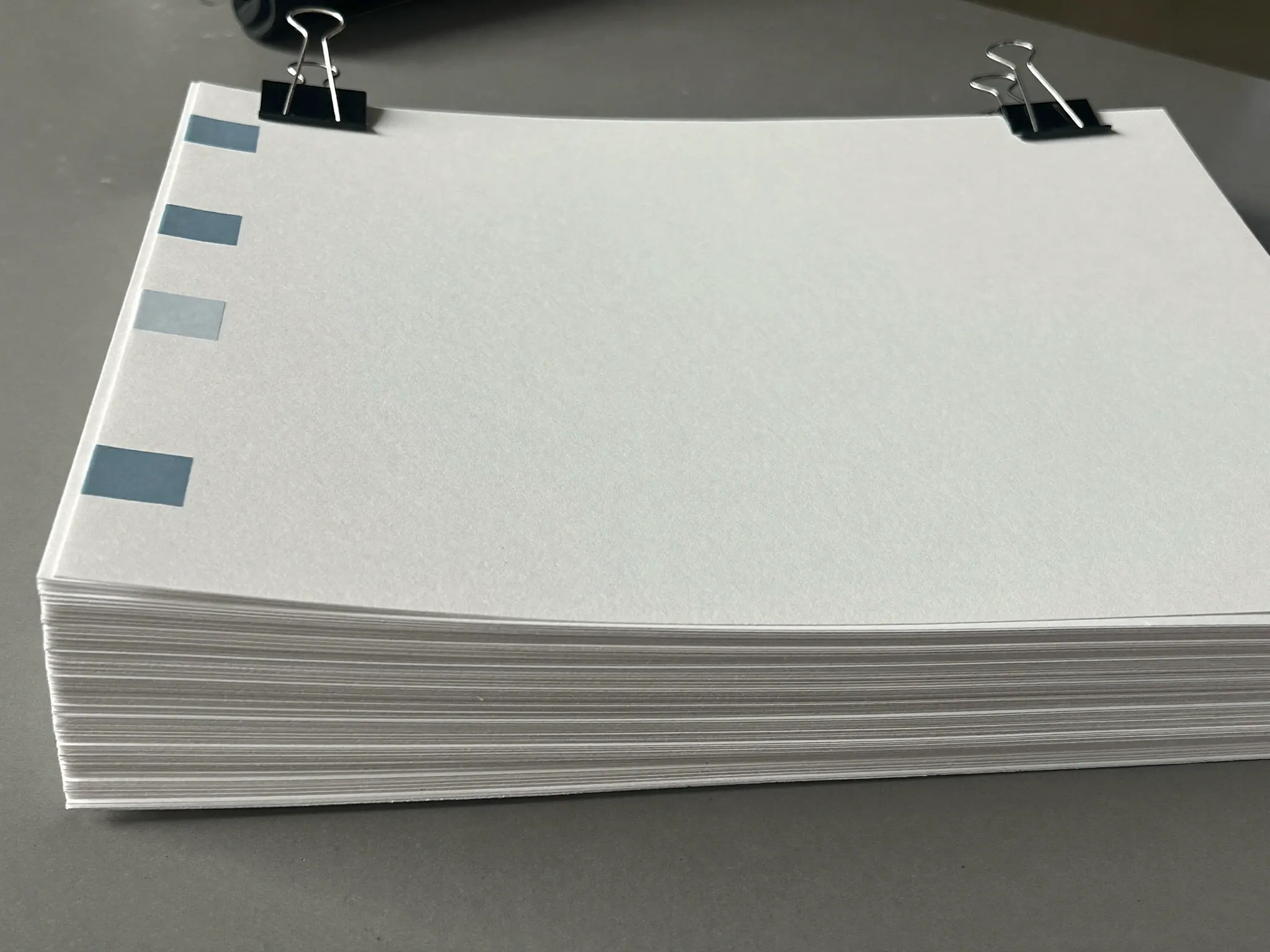 A thick stack of blank white paper with pale blue index tabs and two black binder clips, on a grey desk