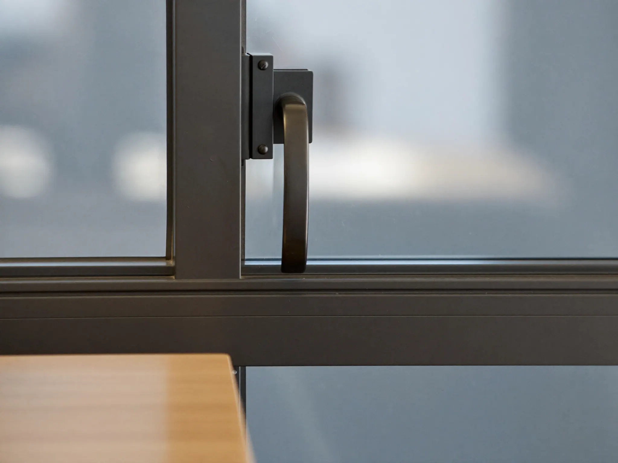 A dark steel window latch and mullion in close-up, with a pale oak desk edge out of focus below