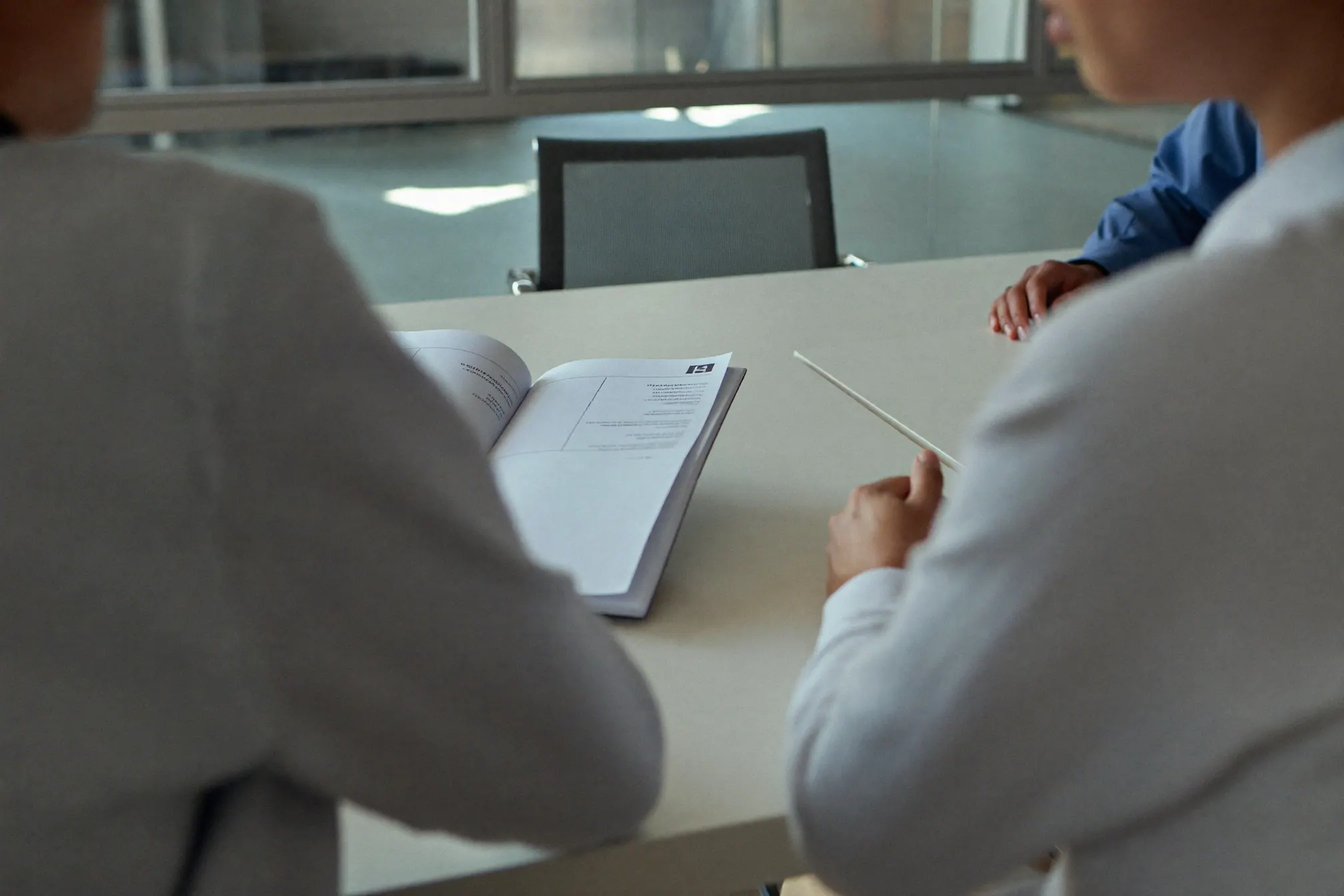 Over the shoulder of two people at a pale meeting table in a glass-walled office, a printed report open between them