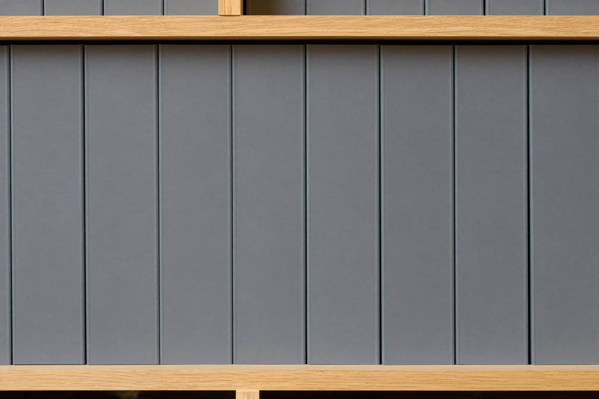 A long row of identical slate-grey document binders on a pale oak shelf in even daylight